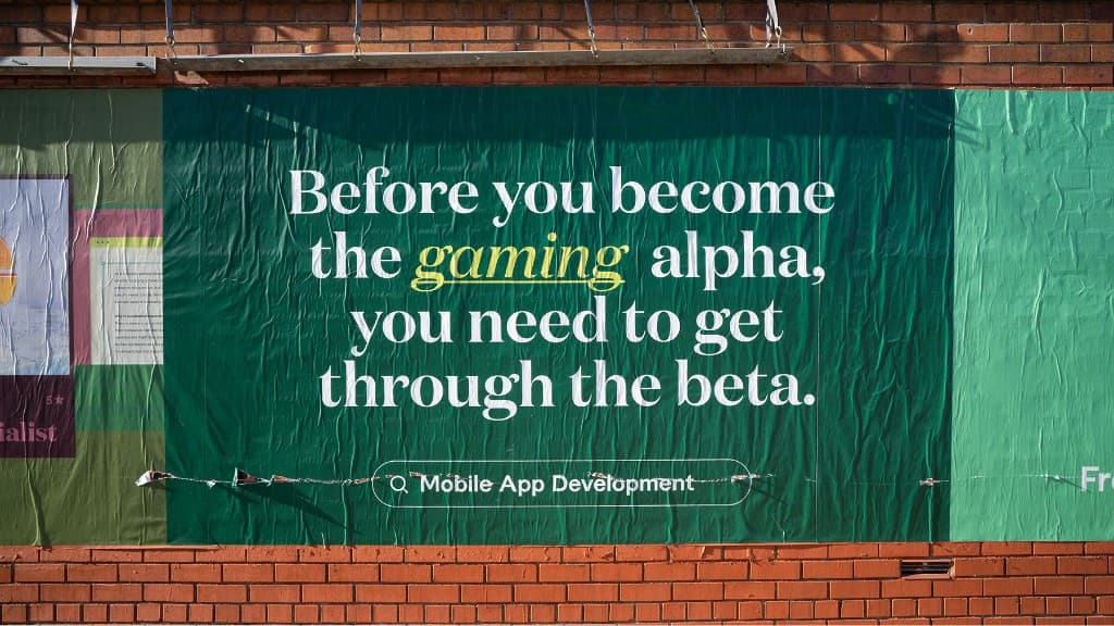 Wheat-pasted poster on brick: Before you become the gaming alpha you need to get through the beta—yellow script gaming, Mobile App Development search pill, mint side stripe