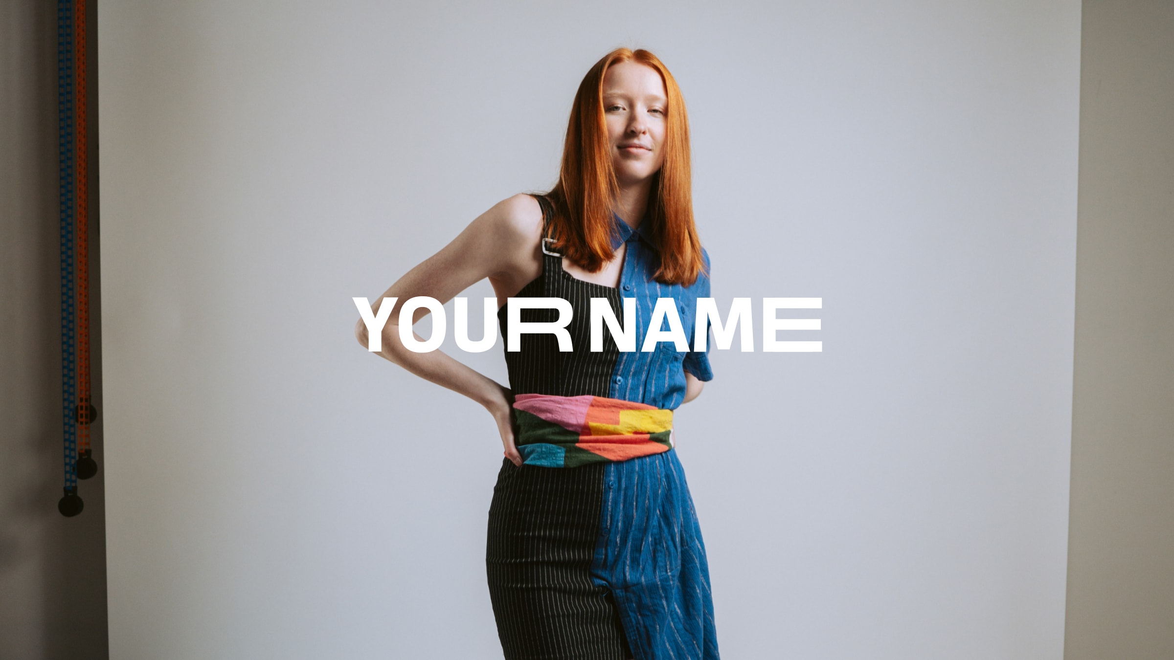 Studio portrait: model with long red hair in a split pinstripe-and-stripe shirt, patchwork sash, large YOUR NAME type across the frame, light gray backdrop