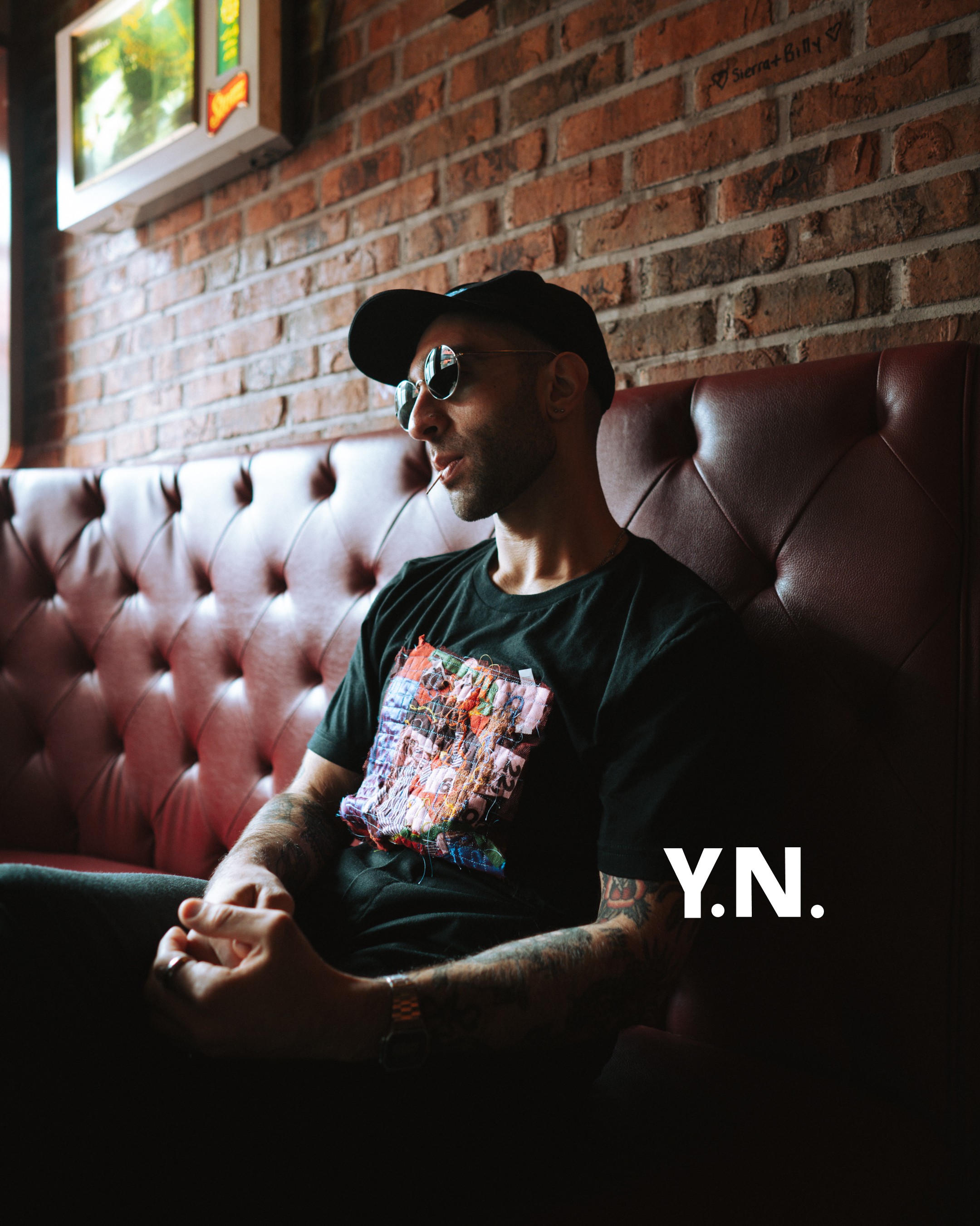Portrait in a red tufted leather booth against brick: cap, sunglasses, graphic patch tee, Y.N. mark, tattoos and watch