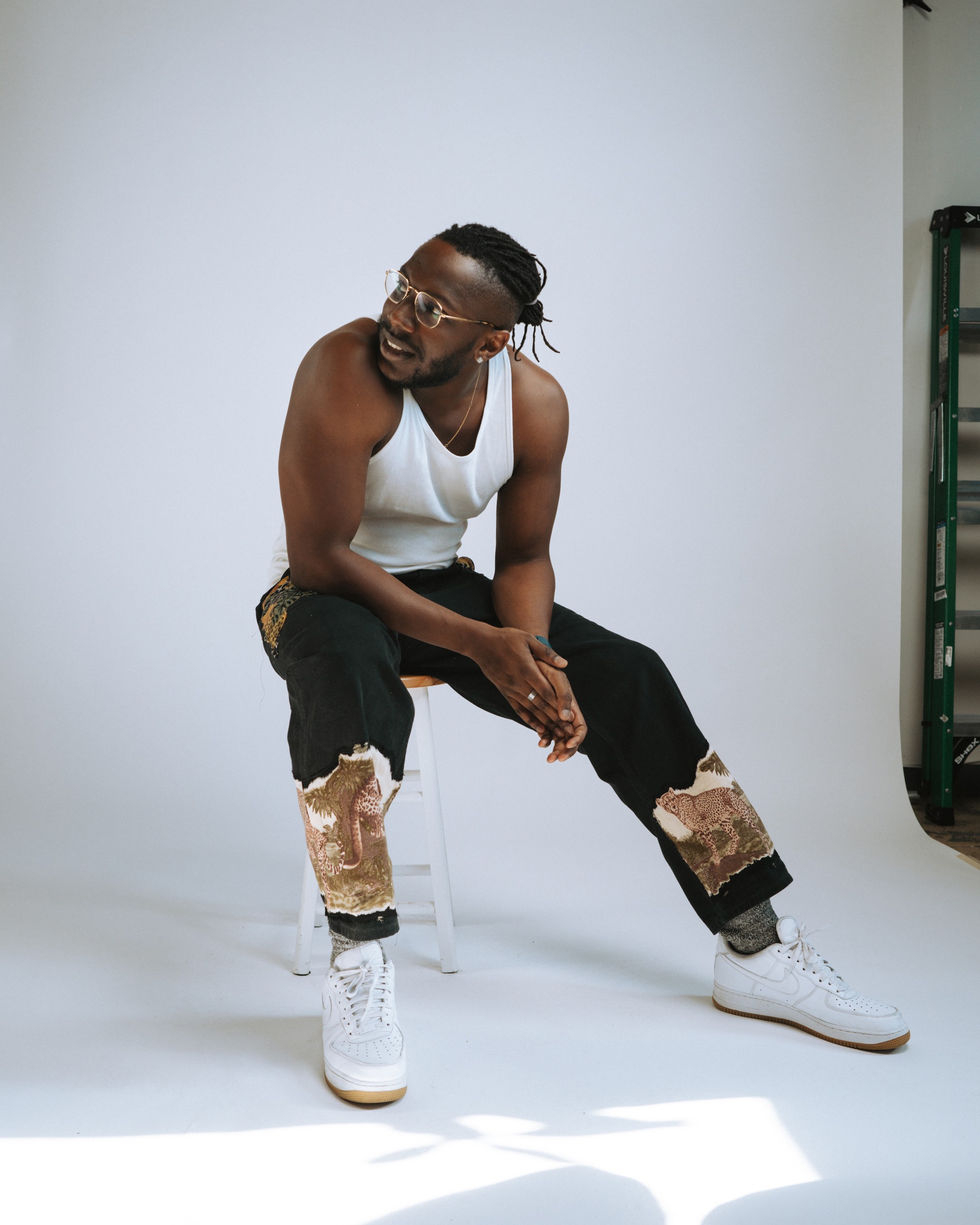Full-length studio portrait on a white stool: white tank, black jeans with large leopard tapestry patches, white sneakers, seamless white floor