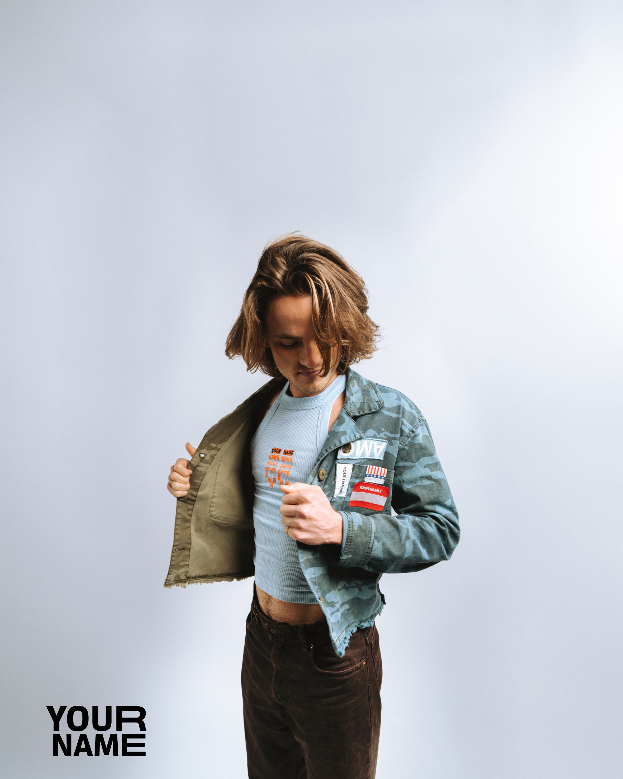 Studio portrait: model opens a camo jacket over a blue ribbed top printed with stacked YOUR NAME type; YOUR NAME lockup in the corner