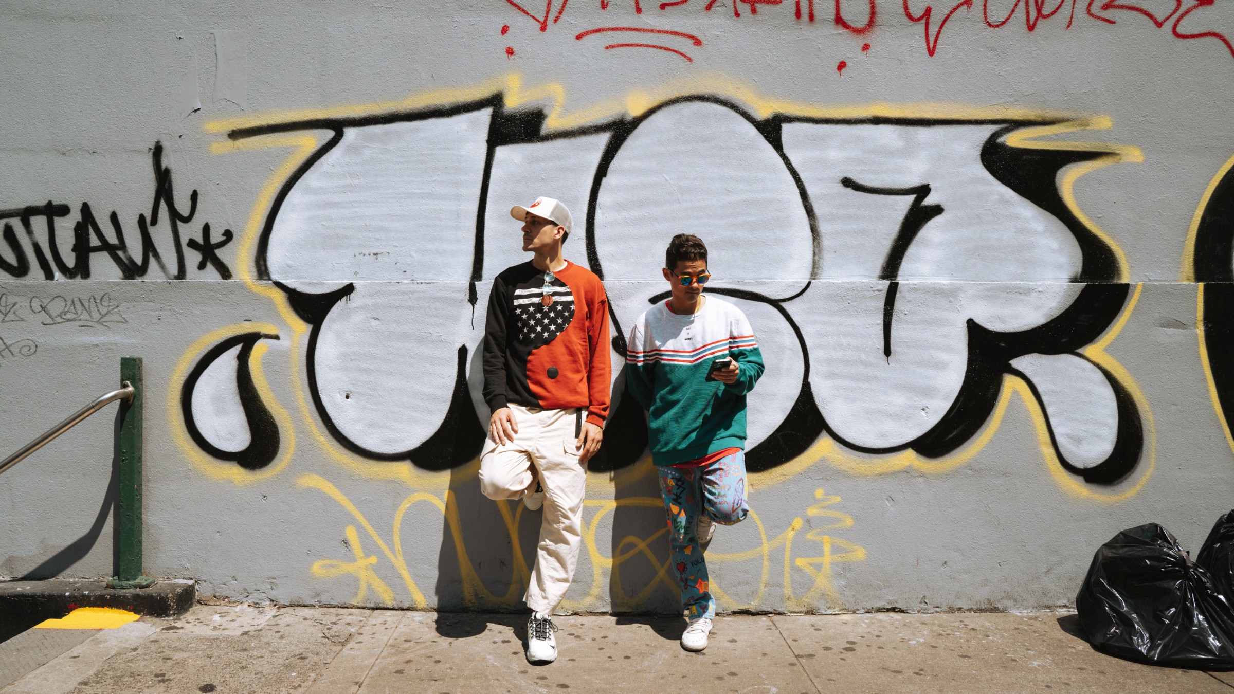 Two models in streetwear against a graffiti-covered wall, bright sun and sidewalk shadows, one checking a phone